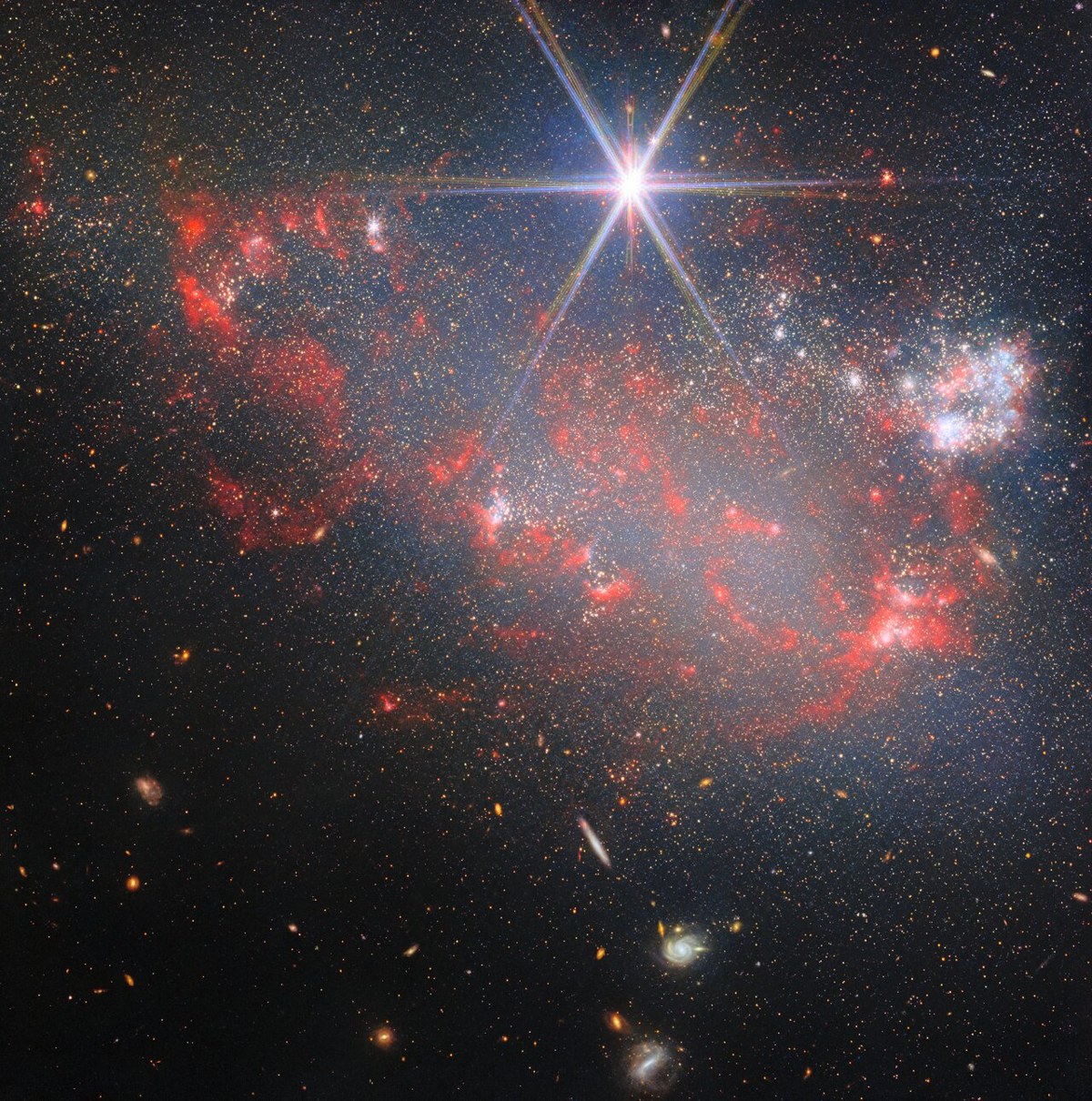 An irregular galaxy, appearing like a broad band of tiny stars. It is denser on one side, with a faint cloud of gas among the stars. Clumps of brighter points are newly formed stars, with the largest lights being star clusters, and with many surrounded in glowing blue gas clouds. Plumes of red dust surround the galaxy. A foreground star appears very large and bright. A few distant galaxies appear in the background.
